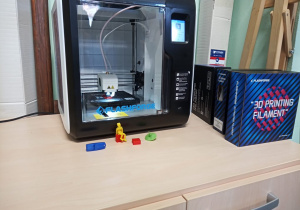 Na zdjęciu znajduje się Drukarka 3D, która stoi na komodzie. Przy drukarce są ułożone figurki, które w niej powstały.