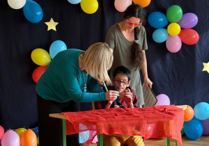 Uczeń siedzi przy stoliku i z pomocą nauczycielek prezentuje swój talent.
