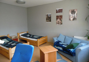 Zdjęcie przedstawia sypialnię. Na pierwszym planie widać miejsce do wypoczynku ładną niebieską kanapę i stolik. W dalszej perspektywie widać dwa drewniane łóżka. Przykryte kocami.